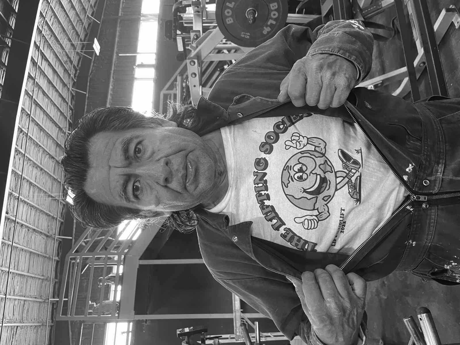 A man in a gym pulling open his jacket to reveal a white t-shirt printed with The Lifting Society logo — a smiling globe mascot giving a thumbs up and flexing, above the tagline 'LIFT AND UPLIFT 2022'.
