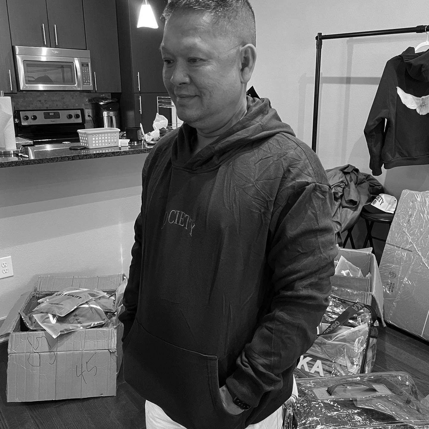 A man smiling in a kitchen wearing a purple Society pullover hoodie, with boxes of inventory at his feet.